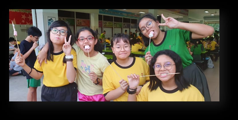 Five children in yellow and green shirts smile and pose with marshmallows on sticks at a school event.